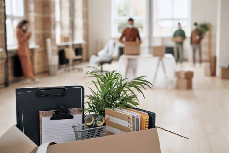 Closeup of opened carton with office items and people settling into the new office in the background in the concept of 'What Are the Factors Influencing the Cost of Office Removals in Greenwich'.